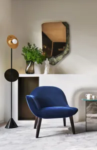 A modern blue velvet armchair in an indoor lounge setting with a unique light fixture, green plant, and abstract mirror.