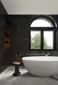 A modern bathroom featuring a dark marble wall, a white freestanding bathtub, and an arched window overlooking green trees.