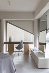 A modern minimalist bedroom featuring a wooden desk, gray chair, and glass table.