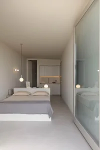 Modern minimalist bedroom with concrete walls and floor, featuring a single bed and a small kitchenette.