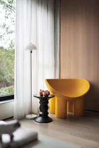 A modern living room featuring a yellow plastic chair, a black side table, and a floor lamp near a window with sheer curtains