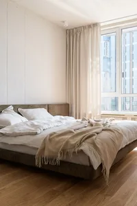 A minimalist bedroom with neutral-toned bedding and curtains, featuring a wooden floor and a concrete-style white wall.