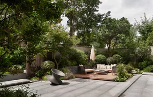 A modern garden featuring wooden seating areas, concrete pathways, and lush greenery surrounded by trees and shrubs.