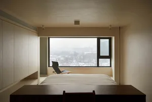 A minimalist bedroom with a large window overlooking a snowy cityscape, featuring a bed, a chair, and a desk.