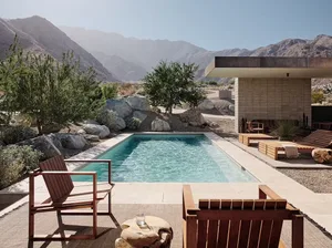 An outdoor living room with a concrete pool, wooden chairs, and surrounding mountainous landscape.