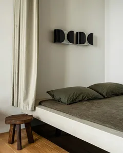A minimalist bedroom featuring a bed with a wooden headboard and green bedding, alongside a wooden stool and a curtain.