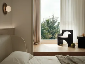 A minimalist bedroom featuring a white bed with a beige headboard, a black armchair with a white cushion, and a window with s