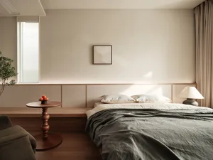 A minimalist bedroom with a neutral color palette, featuring a low-profile bed, a side table with apples, and a window with b