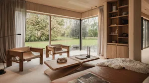 Modern living room with wooden furniture, large windows showing greenery, and a cozy atmosphere.