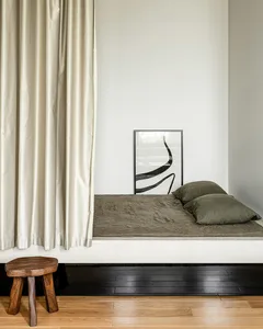 A minimalist bedroom featuring a simple bed with dark bedding, a wooden stool, and a framed artwork on the wall.