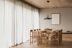A modern dining room with large windows, natural light, wooden furniture, and a minimalist light fixture.