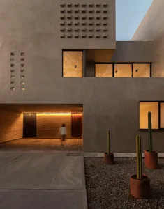 A minimalist concrete house with a person walking through the entrance, featuring several windows and a cactus garden.