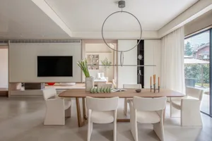 Modern minimalist dining room with white chairs and a wooden table, featuring a round hanging light fixture and a TV in the b