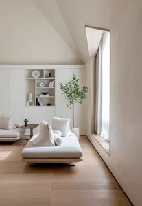 A minimalistic residential interior with neutral tones featuring a daybed, bookshelf, and window