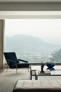 Modern living room featuring a glass coffee table, plush carpet, and a velvet armchair by a large window.