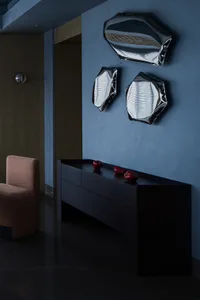 A set of three minimalistic, metallic decorative lights mounted on a blue wall, with a dark wooden cabinet and a brown uphols