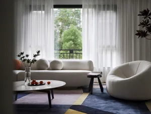 A minimalist living room with white furniture, a blue and yellow rug, and sheer curtains. The room features a low-profile sof