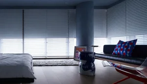 A minimalist living room with soft furnishings, featuring a black leather sofa, red polka dot pillow, and white pleated blind