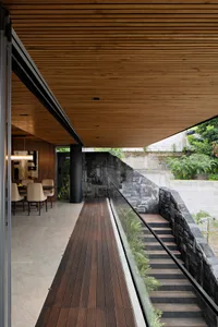 Modern residential exterior with wooden deck, stone wall, and glass railings.