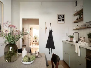 A minimalist kitchen with wood accents features a table with flowers, green fruit, and a white pitcher.