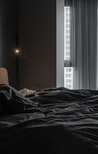 A minimalist bedroom with a black bed and fabric bedding, positioned near a window with sheer curtains and an adjacent floor