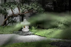 A minimalist rock garden featuring a stone lantern and a small tree, surrounded by green plants and white gravel.