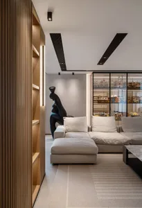 A modern, minimalist living room with a beige sectional sofa, a black sculpture, and a glass cabinet displaying various objec