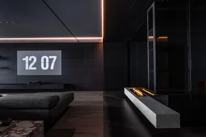 A modern minimalist living space featuring a concrete fireplace and black walls with a clock displaying the time 12:07.
