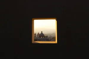 A square window frame reveals a view of a cityscape with domed structures and mountains in the distance, illuminated by a gol