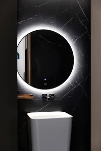 Modern bathroom with a sleek, round mirror with backlit LED lighting and a white ceramic pedestal sink against a black marble