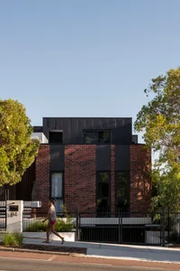 A modern residential house with a brick facade and black accents, set against a clear blue sky with lush green trees surround