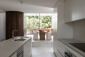 A modern kitchen with a dining table in the center, featuring white cabinetry, a wooden dining table, and glass front cabinet