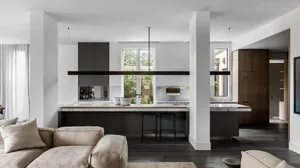Modern kitchen with marble countertops and wooden cabinets, featuring metal stools and a view of the outdoor garden.