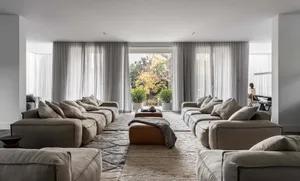 A modern living room with grey fabric sofas arranged in a U-shape, surrounded by large windows with sheer curtains, and featu