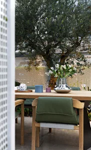 Outdoor dining area featuring a wooden table, green cushioned chairs, and a vase of white flowers.