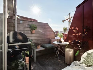Urban rooftop space featuring a wooden bench, metal grill, and potted plants.