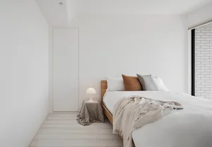 A minimalist bedroom with a white bed, wooden frame, and a bedside lamp on a beige fabric-covered table.