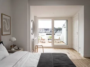 A minimalist bedroom with a wooden floor, white bedding, and a view of a balcony with chairs.