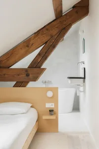 A minimalist bedroom with white walls, a wooden bed frame with a white mattress and pillows, and wooden beams on the ceiling.