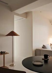 A minimalist living room with a neutral color palette, featuring a sofa, a table with a plate and mug, and a lamp with a coni