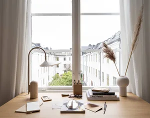 A modern desk setup featuring a white vase, lamp, and books against a window with a view of buildings.