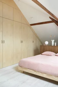 Modern minimalist bedroom featuring a wood platform bed with pink bedding, situated in a slanted ceiling room with exposed wo