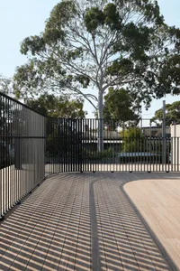 A modern minimalist outdoor patio with wooden flooring and black metal railings, surrounded by trees and natural light.