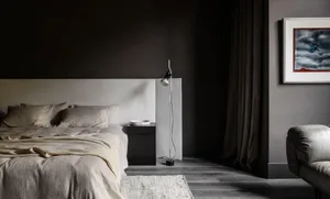 A minimalist bedroom with neutral textiles and a concrete bed frame, featuring blinds and a floor lamp.
