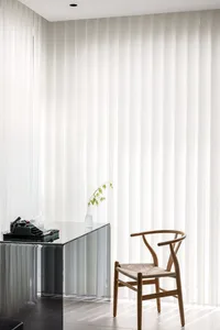 A minimalist desk space featuring a glass desk and a wooden chair with a single stem of greenery.