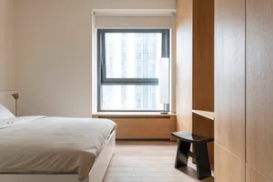 A minimalist bedroom with a white bed, a built-in wooden wardrobe, and a window with a city view.