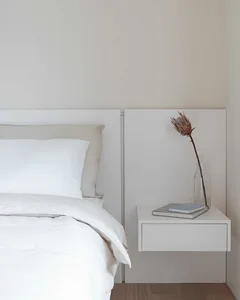 A serene, minimalist bedroom featuring a white bed with neatly arranged white bedding and a simple, white nightstand with a v
