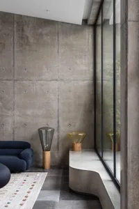 A modern minimalist living room featuring a dark blue sofa, a curved concrete bench, and geometric floor lamp on a patterned 