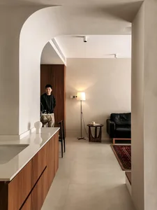 A minimalist living room featuring a concrete floor, a black leather sofa, and a small wooden side table with a lamp and a cu