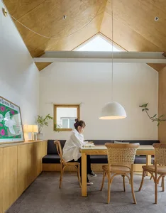A minimalist living room with wooden furniture and a white wall, featuring a woman sitting at a wooden table.